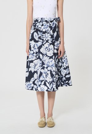 Femme portant une jupe portefeuille bleu marine à grande imprimé floral blanc, associée à des chaussures beige à lacets, debout devant un fond uni.