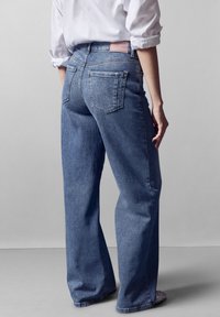 Jeans anchos de mezclilla de color azul claro con bolsillos traseros y una etiqueta rosa en la cintura. Textura suave y ajuste relajado.