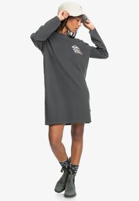 Grijze oversized sweater jurk met lange mouwen, voorzien van een kleine grafische print, gedragen met zwarte gestippelde sokken en groene enkellaarsjes.