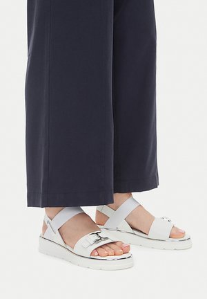 Weiße Plateau-Sandalen mit silbernen Schnallen und grauen Knöchelriemen, mit einer glatten Oberfläche und einer dicken Sohle, kombiniert mit dunkler Hose.