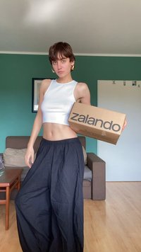 Zalando škatla v levi roki; oblečen v belo brezrokavno majico in ohlapne črne hlače; minimalističen dizajn; notranja postavitev.
