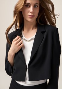 Blazer nero accorciato realizzato in tessuto liscio, con una semplice chiusura, indossato sopra una maglietta bianca con una collana scintillante, che mette in risalto l'eleganza.