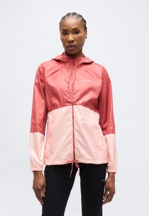 Naine seisab kahetoonilise roosa Columbia kapuutsiga jope ja mustade pükstega ühevärvilise tausta ees.