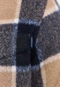 Brown and navy plaid fabric with a soft texture, featuring a small blue label with "Mustang" stitched on the edge.