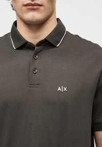 Homme portant un polo gris foncé avec un bord blanc sur le col et un logo "A|X" brodé sur la poitrine.