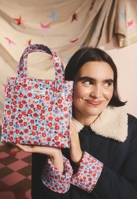 Fiatal nő sötét kabátban, virágos mandzsettával, aki egy virágos mintájú, piros és lila virágokkal és narancssárga csillagokkal díszített táskát tart.