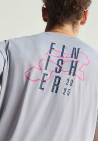 Osoba nosi svijetlosivu majicu s natpisom "FINISHER 2025" i ružičastim apstraktnim linijskim dizajnom na stražnjoj strani, s vidljivim vratom i kovrčavom kosom.