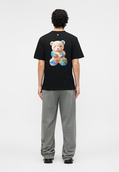 Άνδρας που φοράει μαύρο μπλουζάκι με μεγάλο γραφικό αρκουδάκι που κρατάει πολύχρωμες αφηρημένες σφαίρες στην πλάτη, συνδυασμένο με γκρι φόρμα.