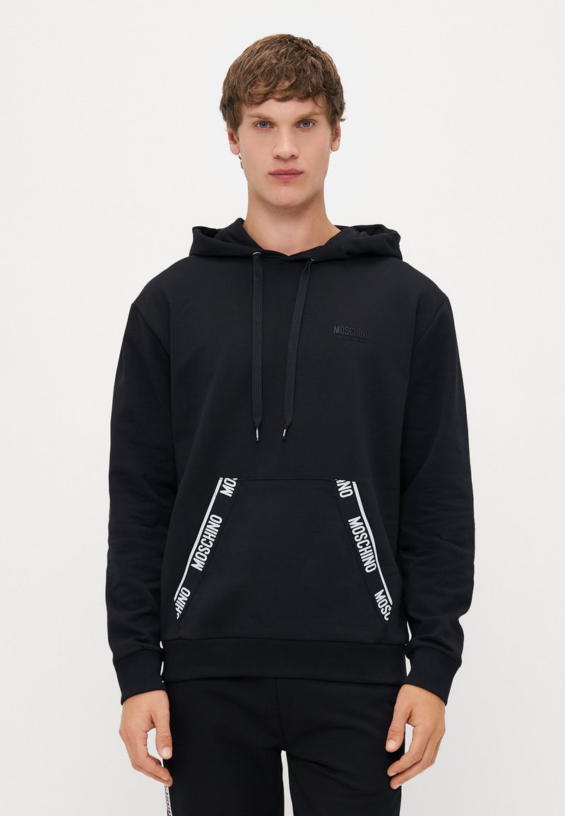 Čierna mikina s kapucňou vyrobená z bavlnenej zmesi. Má predné vrecko s bielym logom Moschino a šnúrky na stiahnutie. Hladká textúra, bežný strih.