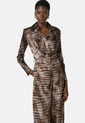 Donna che indossa una camicia marrone e beige a fantasia a maniche lunghe con bottoni e pantaloni a vita alta abbinati con cintura, posando con una mano in tasca.