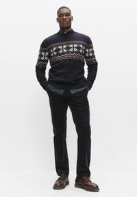 Granatowy sweter z białym i bordowym geometrycznym wzorem, zestawiony z ciemnymi spodniami i brązowymi, sznurowanymi butami. Model stoi z rękami w kieszeniach.