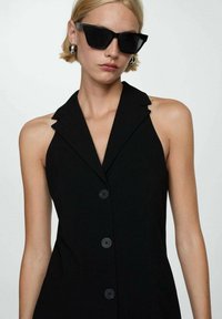 Sort udenærmet kjole med en klippet krave og fem knapper ned ad forsiden. Model med store sorte solbriller og sølvfarvede hoops.