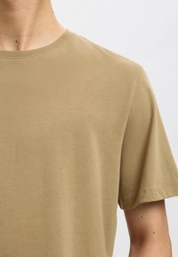 Nærbillede af person iført en ensfarvet beige kortærmet t-shirt med rund hals mod en lys baggrund.
