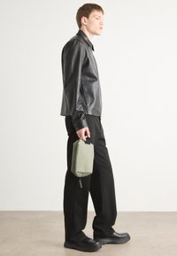 Black leather jacket, loose black pants, and black chunky shoes. Holding a light green rectangular bag with a zip and handle.