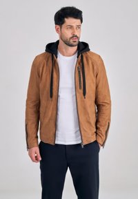 Uomo con capelli scuri che indossa una giacca di pelle marrone chiaro con cappuccio nero, maglietta bianca e pantaloni scuri, in piedi davanti a uno sfondo grigio uniforme.