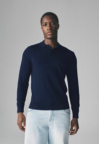 Dunkelblaues Langarm-Polo-Sweatshirt mit Kragen, gerippten Bündchen und Saum. Getragen mit hellblauen Jeans, auf einfarbigem Hintergrund.
