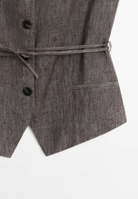 Gilet gris texturé avec des boutons noirs, une fine ceinture nouée et une petite poche avant sur fond blanc.