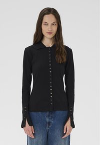 Žena s dlhými hnedými vlasmi, oblečená v priliehavom čiernom top s gombíkmi a predĺženými manžetami a modrých džínoch, stojí na jednostavnom pozadí.