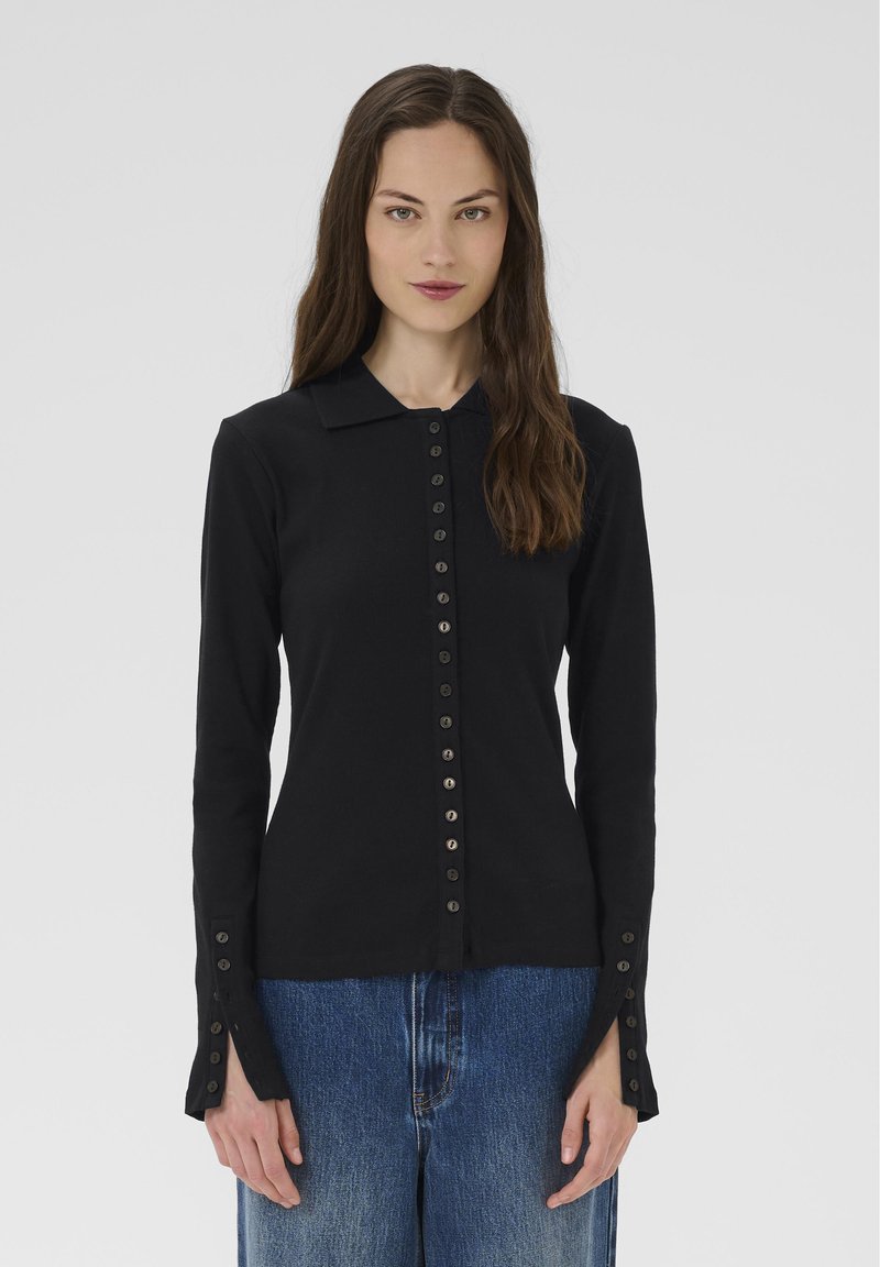 Žena s dlhými hnedými vlasmi, oblečená v priliehavom čiernom top s gombíkmi a predĺženými manžetami a modrých džínoch, stojí na jednostavnom pozadí.