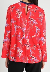 Rote Bluse mit floralem Muster aus leichtem Material, langärmlig, mit einem Schlitz im Rücken, lila- und weißblühenden Muster und einem entspannten Schnitt.