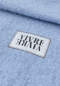 Bílá obdélníková textilní etiketa s černým textem "VIVRE" přišitá na měkkém, světle modrém texturovaném vlněném nebo fleecovém materiálu.