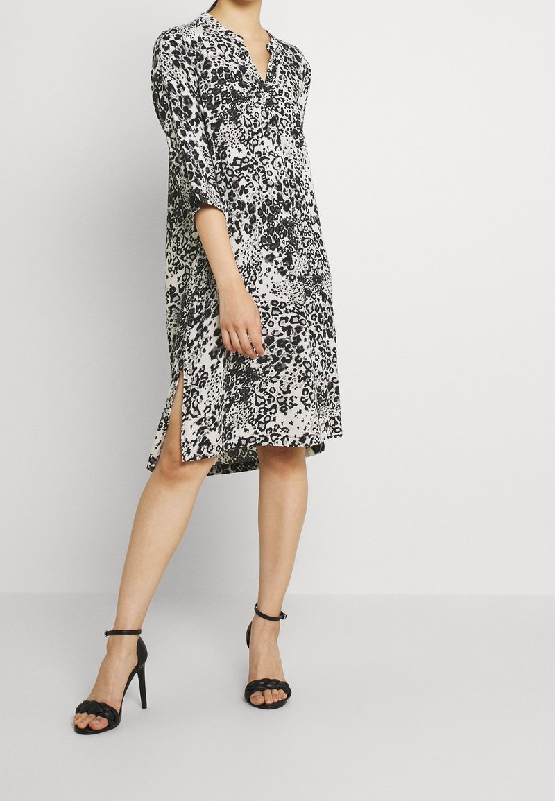 Robe en imprimé léopard avec des manches trois-quarts, un col en V, des fentes sur les côtés et une coupe décontractée. Le tissu semble léger et lisse. Portée avec des talons noirs.