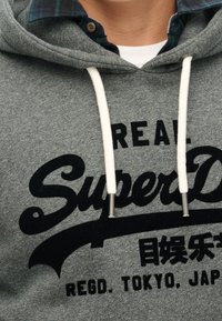 Sudadera gris de algodón con cordones blancos y puntas metálicas. Presenta texto y detalles impresos en negro, sobrepuesta a una camisa de cuadros.