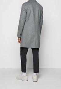 Persona che indossa un cappotto grigio di lunghezza intermedia, pantaloni neri a 3/4, calze bianche e sneakers chunky beige, in piedi contro uno sfondo bianco semplice.