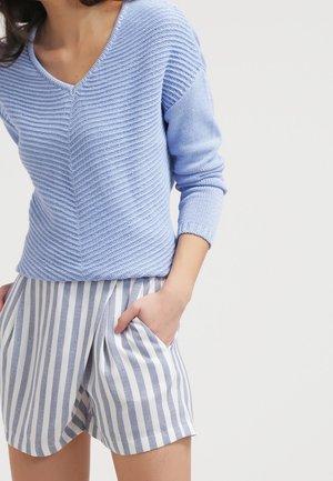 Persoon draagt lichtblauwe gestructureerde V-hals trui en hoog getailleerde witte korte broek met verticale grijze strepen, met één hand in de zak.