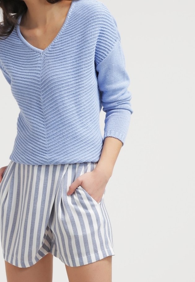 Personne portant un pull texturé bleu clair col en V et un short blanc taille haute avec des rayures grises verticales, une main dans la poche.