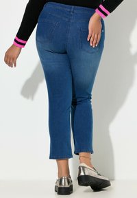 Modré džíny z denimu s přiléhavým střihem a dvěma zadními kapsami, nošené s metalickými platformovými botami a černým svetrem s růžovými pruhy.