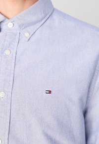Lichtblauw overhemd met knoopsluiting en een button-down kraag, met een klein geborduurd logo in rood, wit en blauw op de borst.