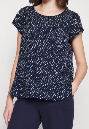 Femme portant un haut à manches courtes bleu marine avec de petits points blancs irréguliers et un pantalon bleu marine assorti, la main dans la poche.