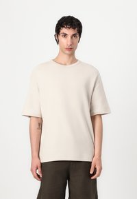 Béžové př oversized tričko s krátkým rukávem, vyrobené z texturované tkaniny, s kulatým výstřihem a volným střihem, nošené s tmavými, volnými šortkami.