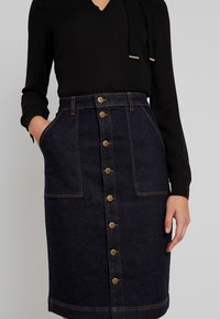 Jeanskjol i mörkblått med hög midja, framfickor och en rad med guldknappar nedanför. Texturerat tyg med kontrasterande sömmar.