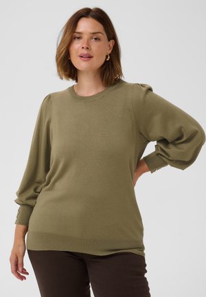 Femme aux cheveux bruns mi-longs porte un pull vert olive à manches bouffantes et un pantalon sombre, debout devant un fond clair et uni.