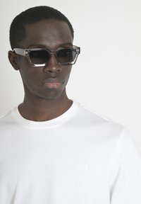 Occhiali da sole quadrati grigi con lenti scure, con dettagli trasparenti, indossati con una t-shirt a girocollo bianca che presenta un logo discreto sul petto.