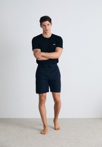 Camiseta negra de cuello redondo con un logo blanco, combinada con pantalones cortos azul marino con cinturilla elástica y cordón, de pie sobre una alfombra beige.