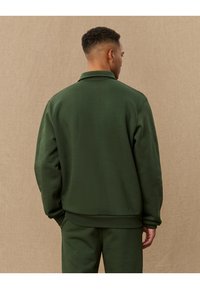 Dark green, collared sweatshirt with ribbed cuffs and hem. Soft, smooth fabric, featuring a relaxed fit and minimalist design.