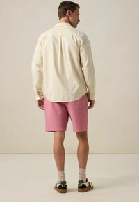 Männliches Outfit mit einem blassgelben gestreiften Hemd und strukturierten rosa Shorts, ergänzt durch beiges Socken und mehrfarbige Sneakers.