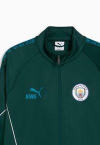 Veste de sport à fermeture éclair vert foncé avec le logo Puma, le mot « KING » et l'écusson du club de football de Manchester City à l'avant.