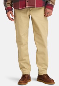 Chinos de algodón beige con corte de pierna recta, que cuentan con bolsillos laterales y una textura suave, combinados con zapatos de cuero marrón.