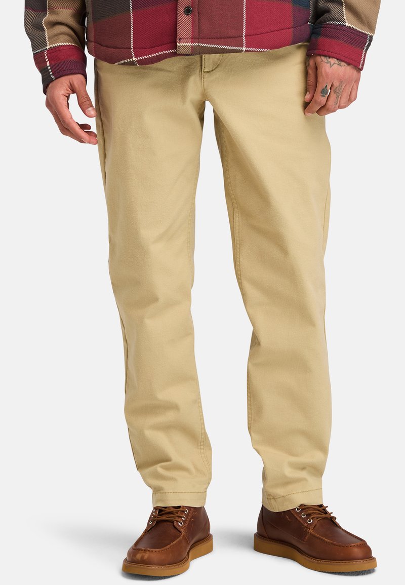 Chinos de algodón beige con corte de pierna recta, que cuentan con bolsillos laterales y una textura suave, combinados con zapatos de cuero marrón.