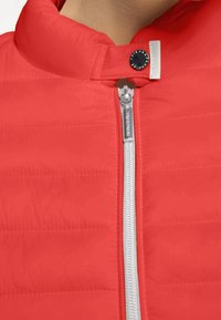 Chaqueta acolchada roja con un cuello alto, que presenta un zipper plateado y un cierre de presión negro. La tela tiene una textura suave con costuras horizontales.