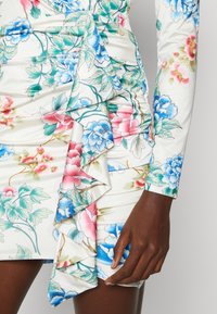 Robe bodycon florale en satin avec une base blanche, présentant des pivoines roses et bleues, un design froncé et un détail de volants superposés à l'ourlet.