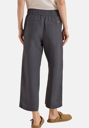 Person trägt lockere dunkelgraue verkürzte Hose mit elastischem Bund und braunen Slippern, steht mit dem Rücken zur Kamera.