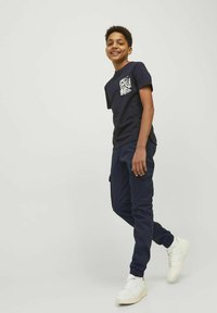T-shirt bleu marine avec une impression de poche florale blanche, associé à un pantalon de jogging bleu foncé et des baskets blanches. Design simple et décontracté.