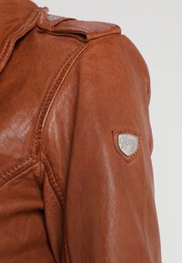 Brun läderjacka med synlig sömnad på axeln och en liten sköldformad metalllogotagg på överarmen.