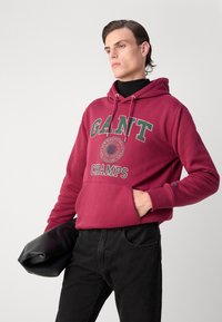 Felpa rossa con cappuccio e grafica "GANT CHAMPS", tasca frontale, jeans neri e borsa a tracolla nera. Tessuto liscio, vestibilità casual.