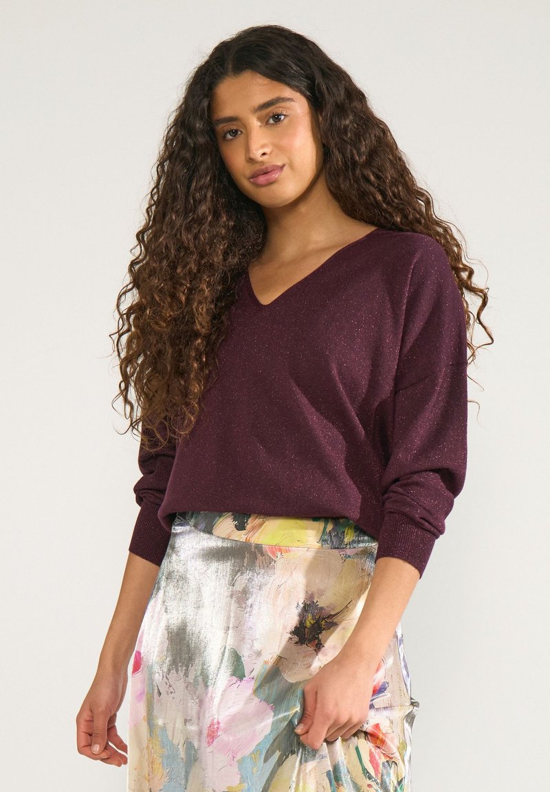 Jeune femme aux longs cheveux bouclés portant un pull violet foncé à col en V et une jupe fleurie brillante, debout devant un fond uni.